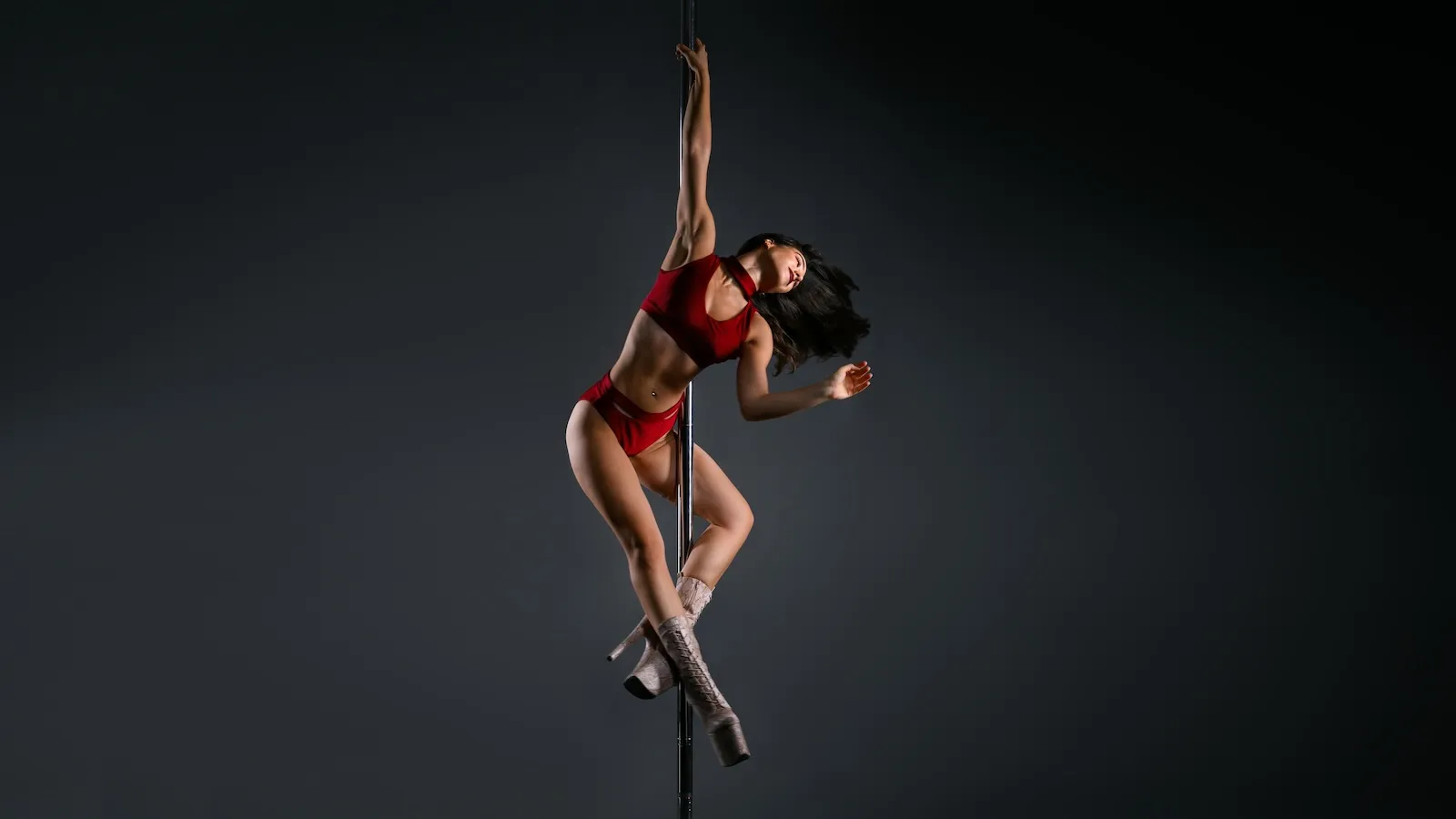 Dancer in a red outfit and high heels, exhibiting strength and dynamism while holding the pole in a powerful pose against a dark background.