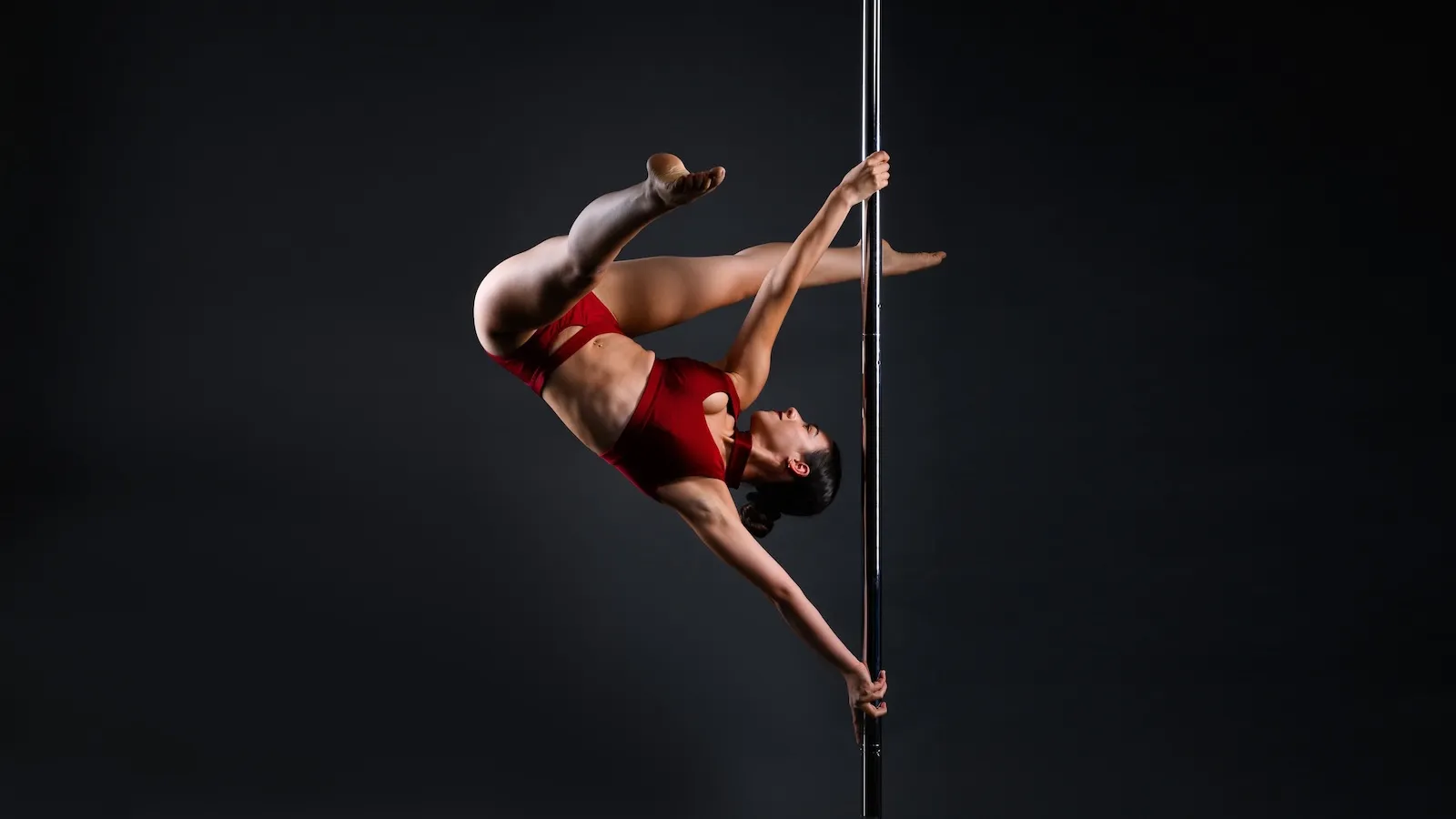 Dancer in a red outfit, showcasing extreme flexibility and control in a back arch pose, holding the pole with both hands and lifting both legs upward.