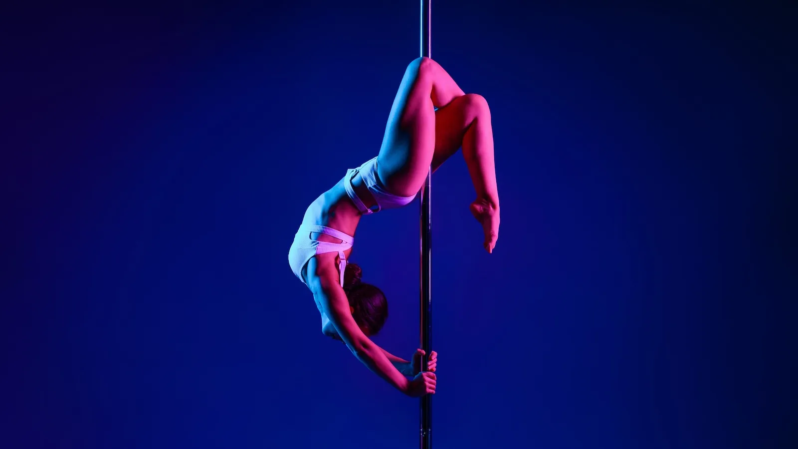 Dancer in a white outfit, sitting gracefully at the base of the pole in a contemplative pose, illuminated by ethereal blue lighting.