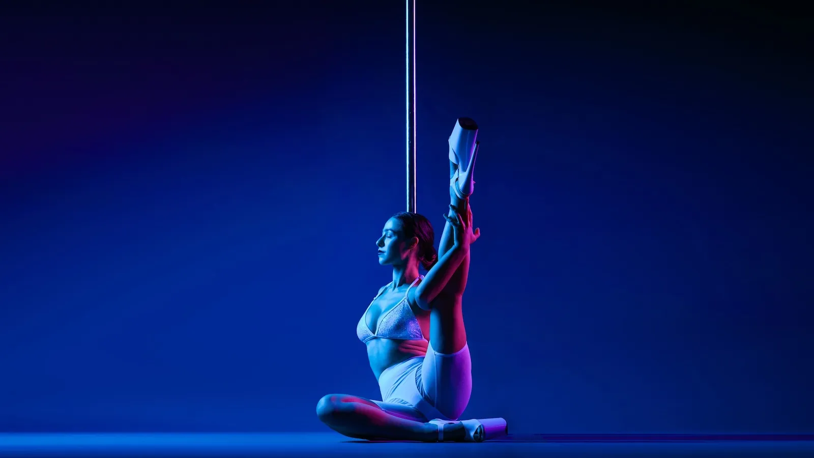 Dancer in a white outfit, performing an inverted move on the pole, demonstrating strength and agility, highlighted by dramatic blue lighting.